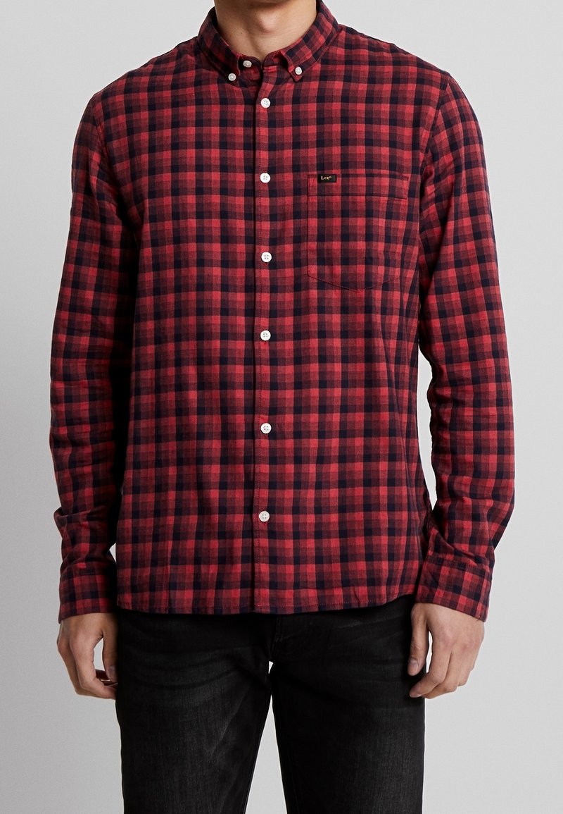 Homme portant une chemise à carreaux rouge et noir avec une poche poitrine et un pantalon noir, debout devant un fond gris uni.