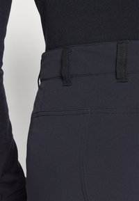 Zwarte stretchstof broek met een gladde textuur, voorzien van riemlussen en zichtbaar stiksels langs de tailleband voor extra detail.
