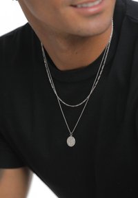 Zilveren gelaagde kettingen op een zwart T-shirt. De ene ketting is glad, de andere is getextureerd en heeft een cirkelvormige hanger met fijne details.