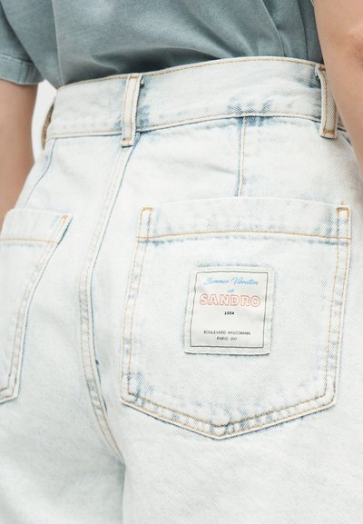 Άτομο που φοράει ανοιχτόχρωμα τζιν με ραφή στην πίσω τσέπη όπου αναγράφεται «SANDRO 1984 Boulevard Haussmann Paris VIII»