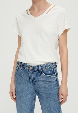 Kobieta w białej koszulce z dekoltem w serek z podwójnymi ramiączkami na ramionach i niebieskich, wysokowzwiązanych dżinsach na jednolitym tle.