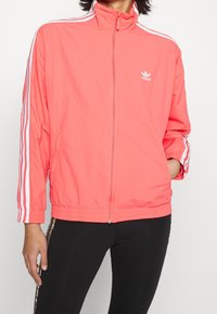 Kvinna som bär en korallrosa Adidas-träningsjacka med vita ränder på ärmarna och dragkedja framtill, tillsammans med svarta byxor.