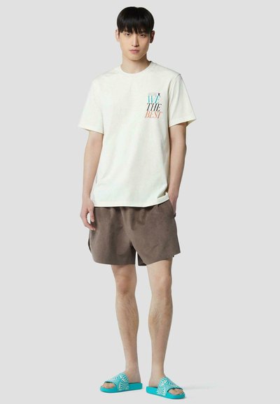 Jeune homme portant un t-shirt blanc graphique, un short marron et des sandales turquoises à enfiler, debout devant un fond clair uni.