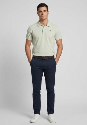 Hombre con camiseta polo verde claro, pantalones azul oscuro, cinturón marrón y zapatillas blancas, de pie con las manos en los bolsillos contra un fondo gris.