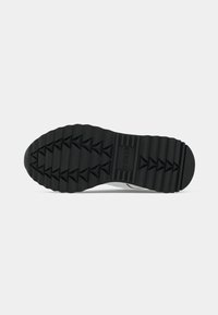 Sole di sneaker in gomma nera con un motivo testurizzato a zigzag e il nome del marchio "GUESS" impresso vicino al tallone.
