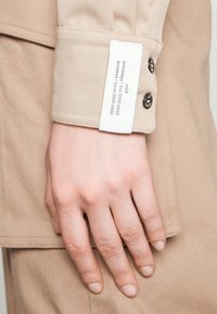 Main reposant à côté d'une manche de veste beige avec deux boutons et une étiquette en tissu blanc indiquant les codes de saison et d'année.