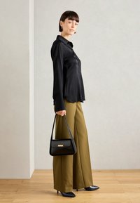 Camicia nera con bottoni, pantaloni larghi color oliva, borsa nera e décolleté a punta, su uno sfondo neutro con pavimento in legno.