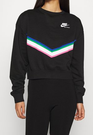 Person iført sort Nike sweatshirt med lyserøde, hvide, grønne og blå chevronstriber på brystet og sorte bukser.
