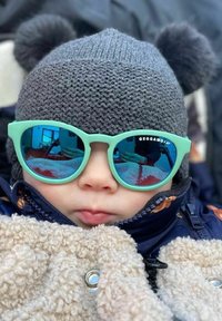 Vaalean teal väriset aurinkolasit peililinssillä, joita käyttä�ä lapsi harmaassa pom-pompulla varustetussa neulepipossa, päällään beige pörröinen takki.