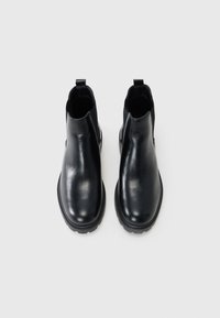 Botas de tobillo de cuero negro con paneles laterales elásticos y lengüetas; textura suave, punta redonda y suelas de goma gruesas.