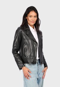 Chaqueta de biker de cuero negra con un diseño recortado, cremallera asimétrica y acentos de cuello con tachuelas, combinada con jeans de mezclilla azul claro.