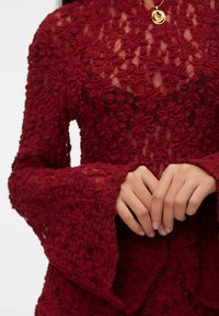 Bordowa koronkowa bluzka o kwiaturowej fakturze, z długimi, rozszerzanymi rękawami i przejrzystym fasonem. Posiada okrągły dekolt i warstwowe zdobienia.