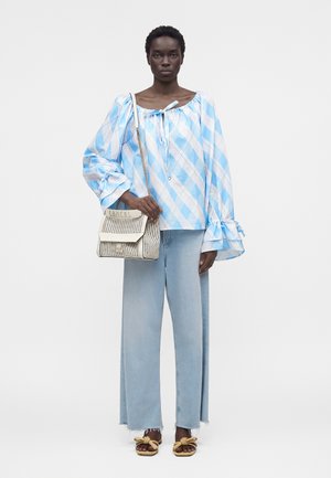 Nainen pukeutunut vaaleansiniseen ja valkoiseen ruudulliseen, pussihihaiseen paitaan, vaaleisiin leveälahkeisiin farkkuihin, ruskeisiin sandaaleihin ja pitää käsissään kermaista punottua käsilaukkua.