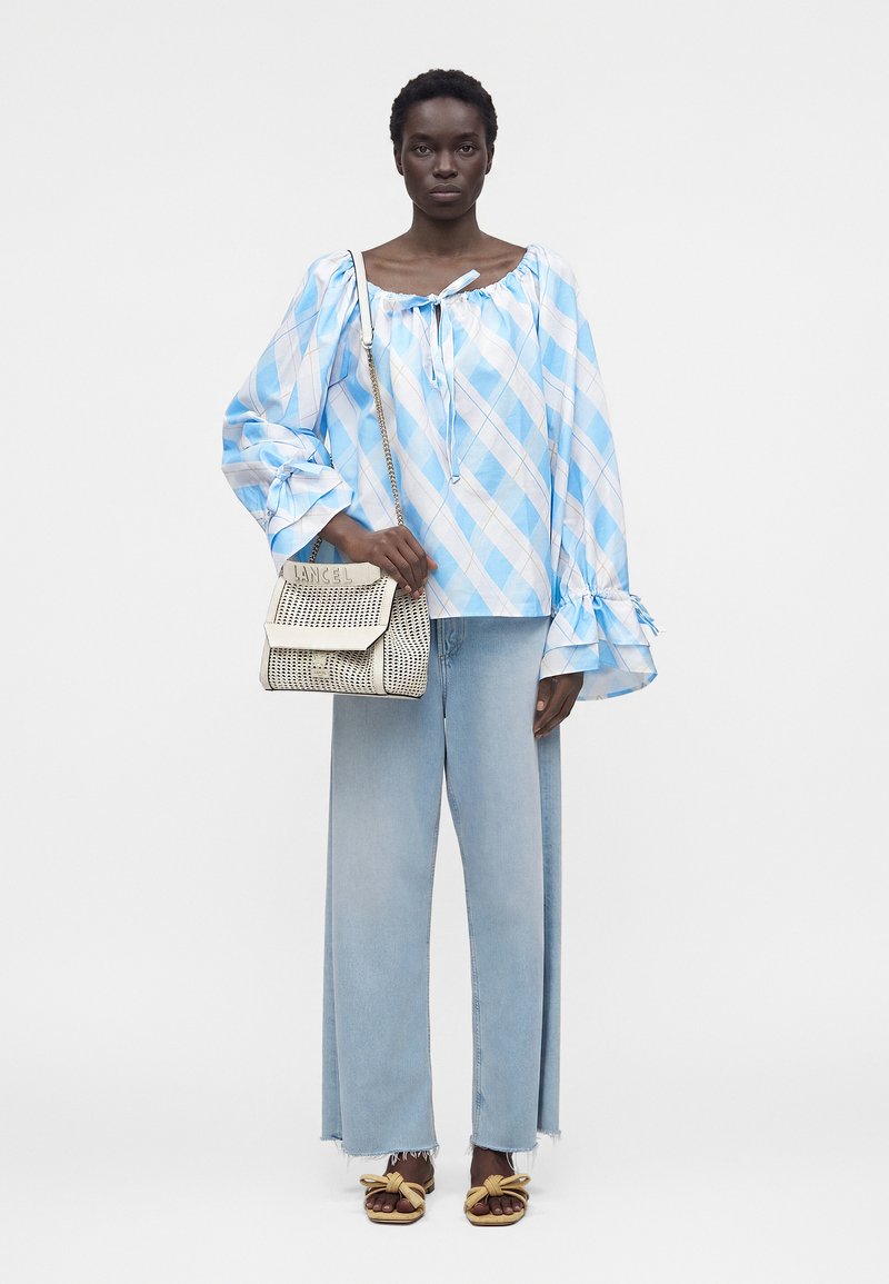 Γυναίκα με ανοιχτογάλανο και λευκό καρό πουκάμισο με φουσκωτά μανίκια, ανοιχτόχρωμα φαρδιά τζιν, καφέ σανδάλια και κρατώντας μια κρεμ πλεκτή τσάντα.