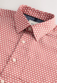 Chemise rouge avec motif floral blanc, col boutonné, texture légère, petits boutons et un accent bleu marine à l'intérieur du col.