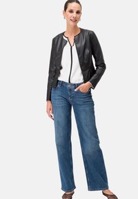 Schwarze Lederjacke mit kurzem Schnitt und rundem Halsausschnitt über einer weißen Bluse, kombiniert mit hellblauen weiten Jeans und schwarzen Netzschuhen.