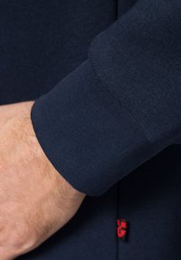 Morska niebieska koszula z długim rękawem ma mankiet z prążkowaną fakturą i gładkim wykończeniem, z małym czerwonym detalem z metką przy szwie.