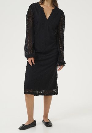 Kobieta w czarnej koronkowej sukience midi z długimi przezroczystymi rękawami, dekoltem w serek i czarnych balerinach, stojąca na jednolitym tle.