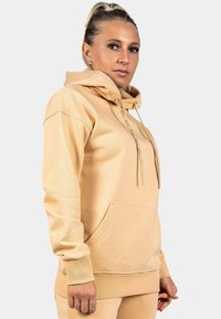 Felpa beige con tasca frontale, cappuccio con lacci e polsini a coste. Il tessuto morbido e la vestibilità rilassata sottolineano il comfort e la praticità.