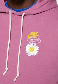 Fialová mikina s kapucí a stahovací šňůrkou, na které je vyšitá bílá sedmikráska a žlutý text "NIKE Z BEAVERTONU S LÁSKOU."