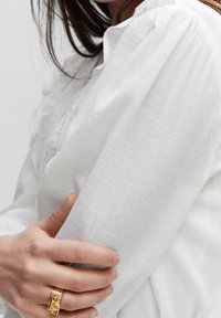 Blouse en lin blanc à manches froncées, avec une texture légère. Une bague en or est portée à la main qui serre la manche.