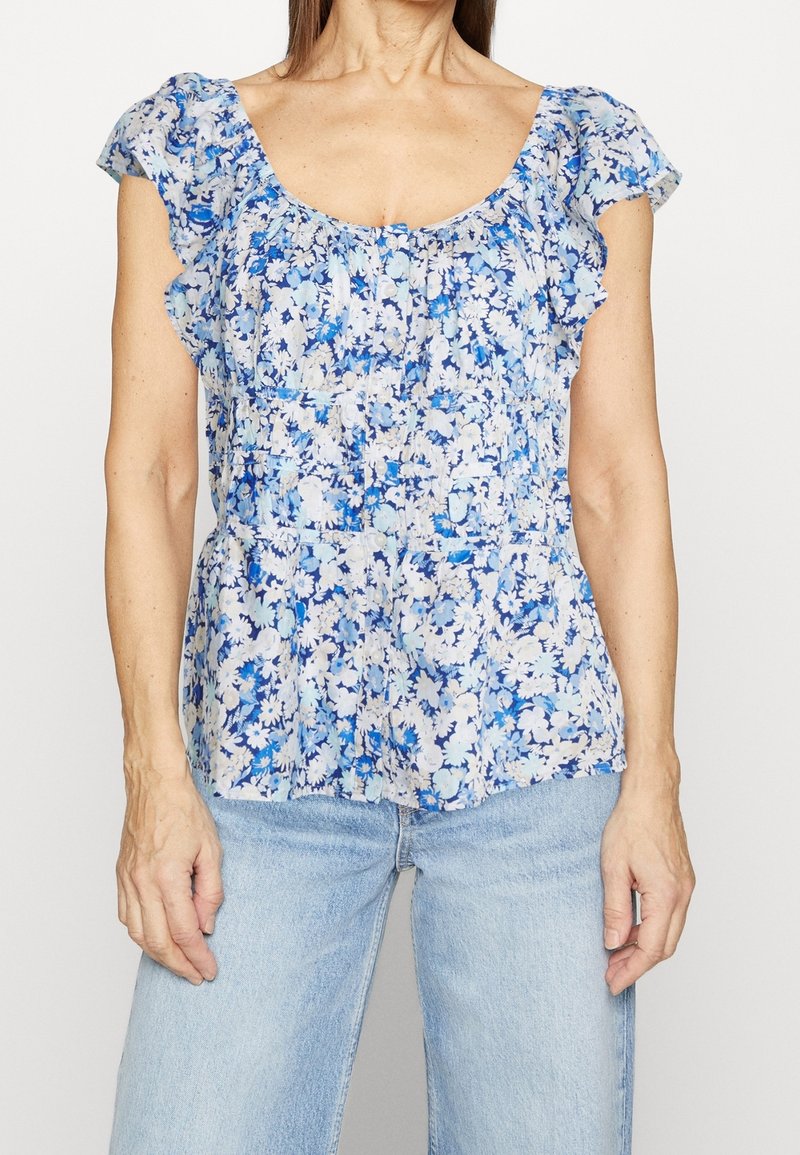 Femme portant un chemisier à manches courtes bleu et blanc à motif floral avec des manches à volants et un jean en denim bleu clair, debout devant un fond uni.