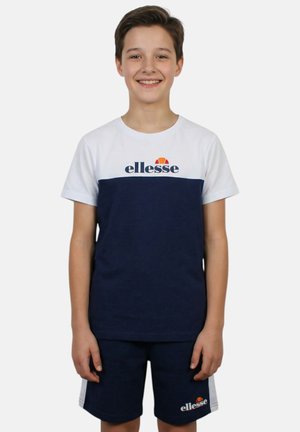 Ragazzo in piedi che sorride, indossa una t-shirt bianca e blu navy ellesse e pantaloncini blu navy abbinati con il logo ellesse.