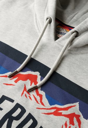 Lichtgrijze hoodie met een trekkoord met metalen tips, opvallende bergafbeelding in rood en blauw, en een groot "SUPERDRY"-logo onderaan.