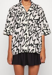 Camicia a maniche corte con bottoni in tessuto bianco, caratterizzata da un audace motivo a onde nere. Dettagli notevoli includono il colletto e una vestibilità rilassata.