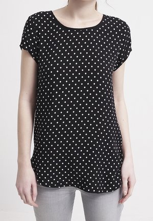 Mujer que lleva una camiseta negra de manga corta con lunares blancos y pantalones gris claro, posando sobre un fondo liso y claro.