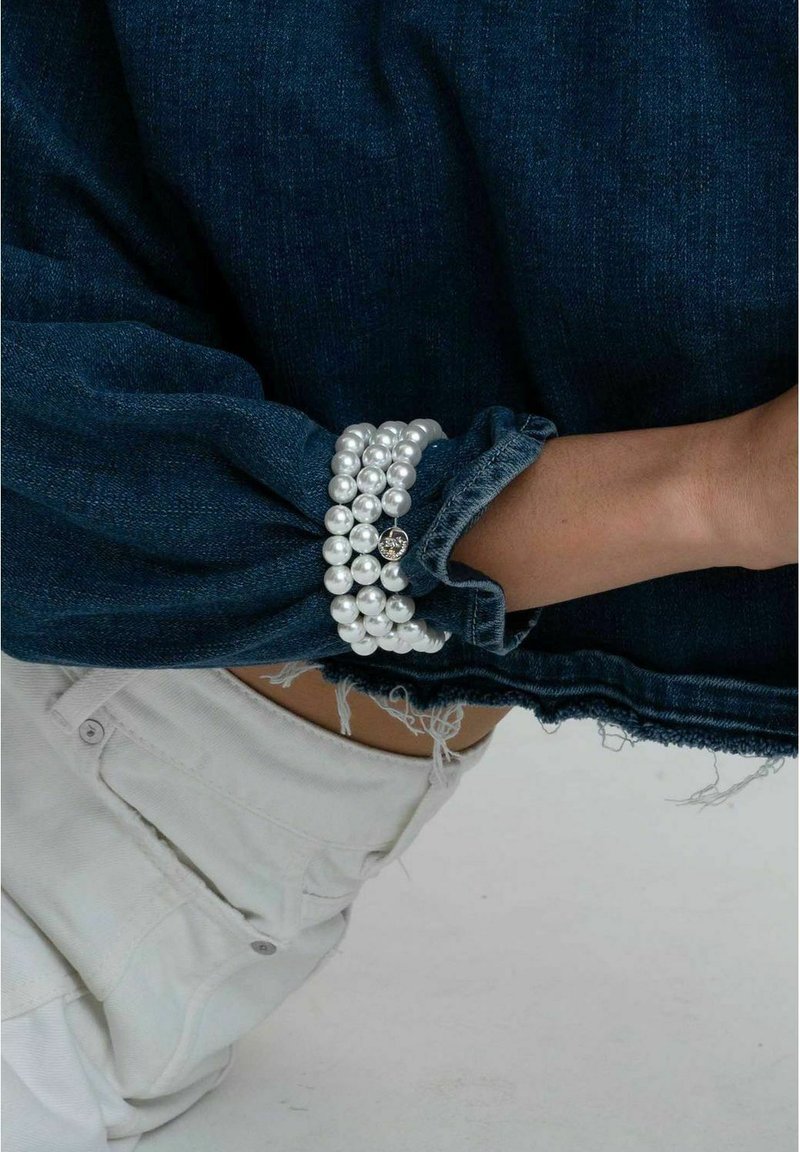 Vitt pärlarmband med en glittrande accent, bärs på en denim cuff. Denimtyget är mörkblått, med en fransad fåll och en något lös passform.