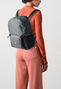 Mochila de tela gris con acentos negros, que cuenta con un bolsillo frontal con cremallera, correas acolchadas y un logotipo de marca en la esquina inferior.