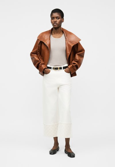 Γυναίκα που φορά καφέ δερμάτινο μπουφάν πάνω από μπεζ κοντομάνικη μπλούζα με ρίγες, λευκό παντελόνι φαρδιάς γραμμής με υφασμένους ρεβέρ, μαύρη ζώνη και μαύρα μπαλαρίνες, όρθια.