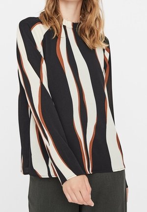 Vrouw draagt een top met lange mouwen met verticale golvende strepen in zwart, wit en bruin, gecombineerd met donkere broek, tegen een effen achtergrond.