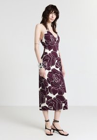 Donna che indossa un abito midi senza maniche con grandi stampe floreali di colore viola scuro, bracciali d'argento e sandali neri con tacco basso e fasce.