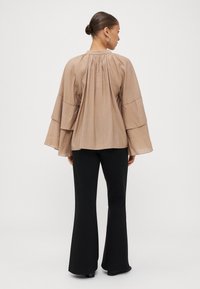 Light brown, long-sleeve top with layered, bell-shaped sleeves and gathered stitching at the yoke. Paired with black flared pants.