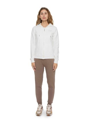 Una mujer está de pie sobre un fondo blanco, con una sudadera blanca con capucha y cremallera, pantalones de chándal color topo y zapatillas deportivas beige.