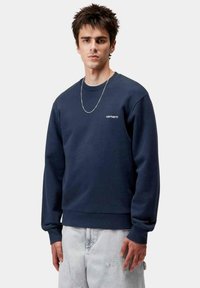 Sudadera de algodón azul marino con cuello redondo y puños acanalados, con un logo blanco de "Carhartt" en el pecho. El modelo lleva unos jeans claros.