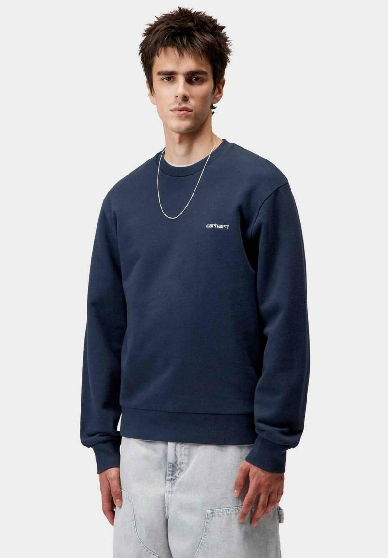 Sudadera de algodón azul marino con cuello redondo y puños acanalados, con un logo blanco de "Carhartt" en el pecho. El modelo lleva unos jeans claros.