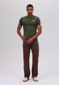 Camiseta verde ajustada con una franja lateral blanca y logo, combinada con pantalones rectos marrones y zapatillas blancas con acentos azules.