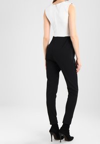 Femme portant un top sans manches blanc rentré dans un pantalon noir taille haute et des bottines à talons noires, se tenant de dos.