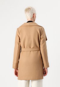 Manteau en laine beige avec une taille ceinturée, un col à revers et des manches longues ; texture lisse et design mi-long.