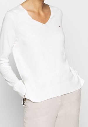 Osoba ubrana w biały sweter z długim rękawem i dekoltem w serek oraz jasnobeżowe spodnie, z rękami swobodnie wsuniętymi do kieszeni, na jednolitym tle.