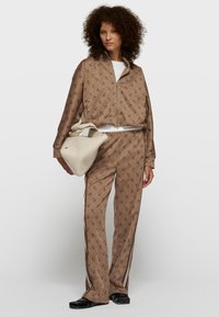 Chándal marrón con patrón y logotipos sutiles, chaqueta con cremallera, cintura elástica, combinado con un bolso crema y zapatos planos negros.