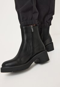 Black leather ankle boots with a side zipper, chunky low heel, and textured sole, paired with black gathered pant cuffs.