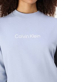 Sweatshirt azul claro feito de algodão, com um decote redondo canelado e texto branco "Calvin Klein" no peito. Tecido liso, sem adornos.