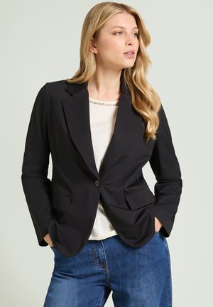 Kvinde iført en sort enkeltknappet blazer over en hvid top og blå jeans, stående med hænderne i lommerne, kiggende let mod højre.