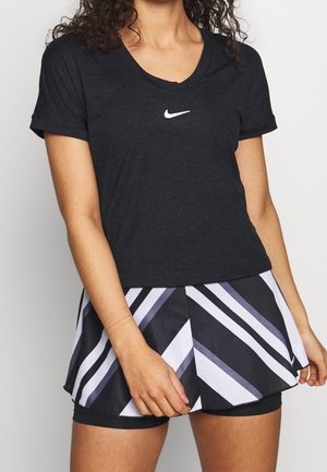 Frau trägt ein schwarzes Nike-T-Shirt und einen schwarz-weiß-lila gestreiften Sportrock mit integriertem Shorts, steht vor weißem Hintergrund.