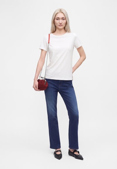 Femme aux cheveux blonds raides portant un t-shirt blanc, un jean bleu, des chaussures noires pointues et tenant un petit sac rouge avec une bandoulière chaîne.