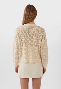 Ljusbeige stickad tröja med ett diamantmönster, vida ärmar och ribbat avslut. Bärs över en krämfärgad kjol.
